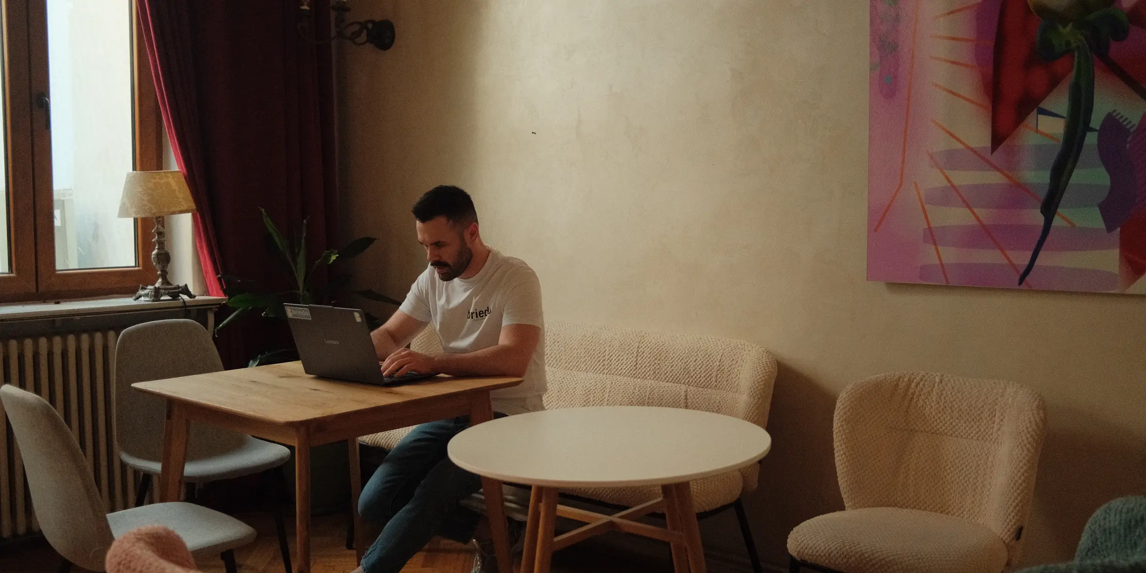 A member working on a laptop at a wooden table in the Nookyard room, with a tall curtained window, soft armchairs and a large abstract painting behind him.